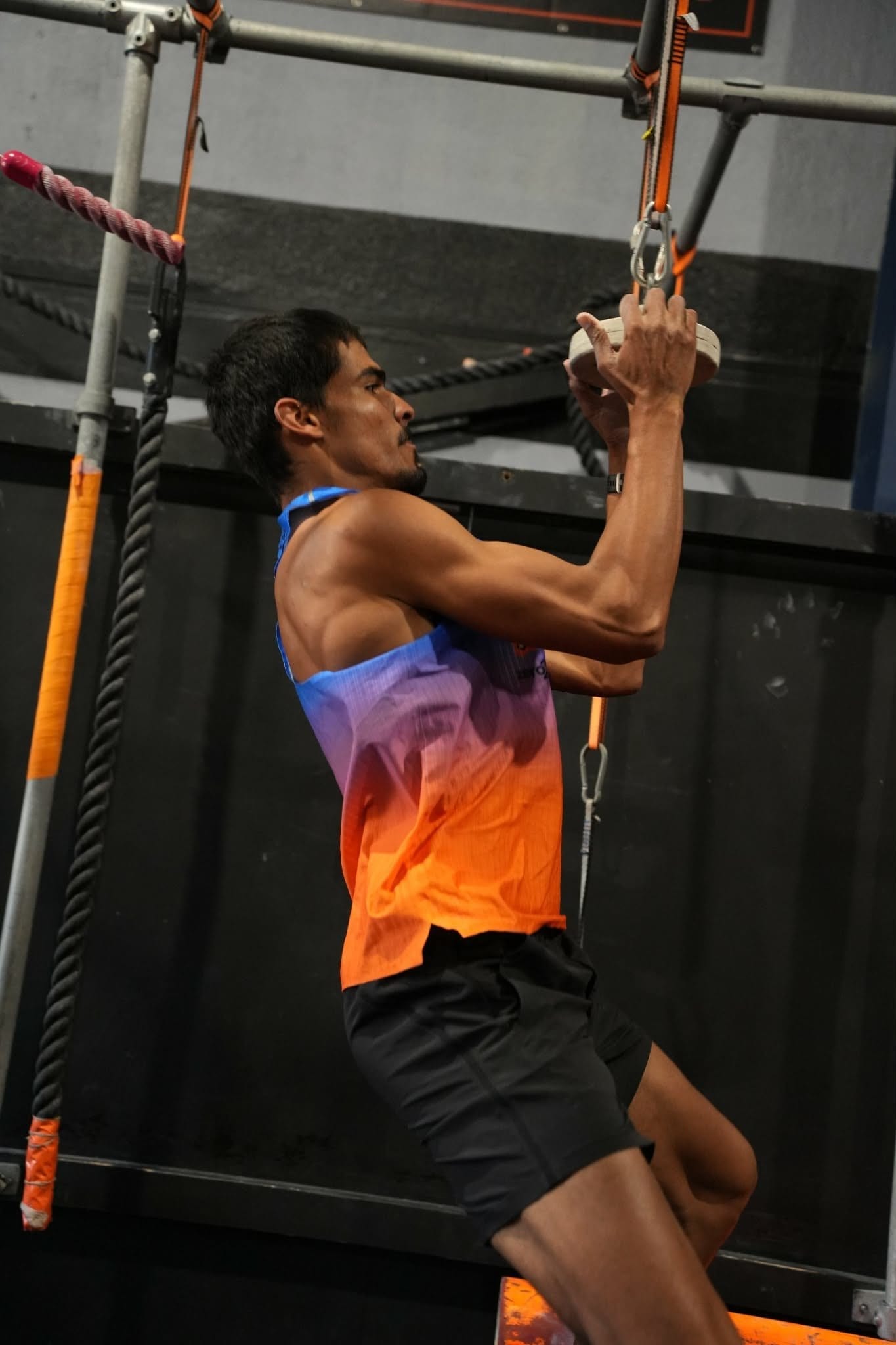 Athlete in an orange singlet hanging from a ring on the AWF rig