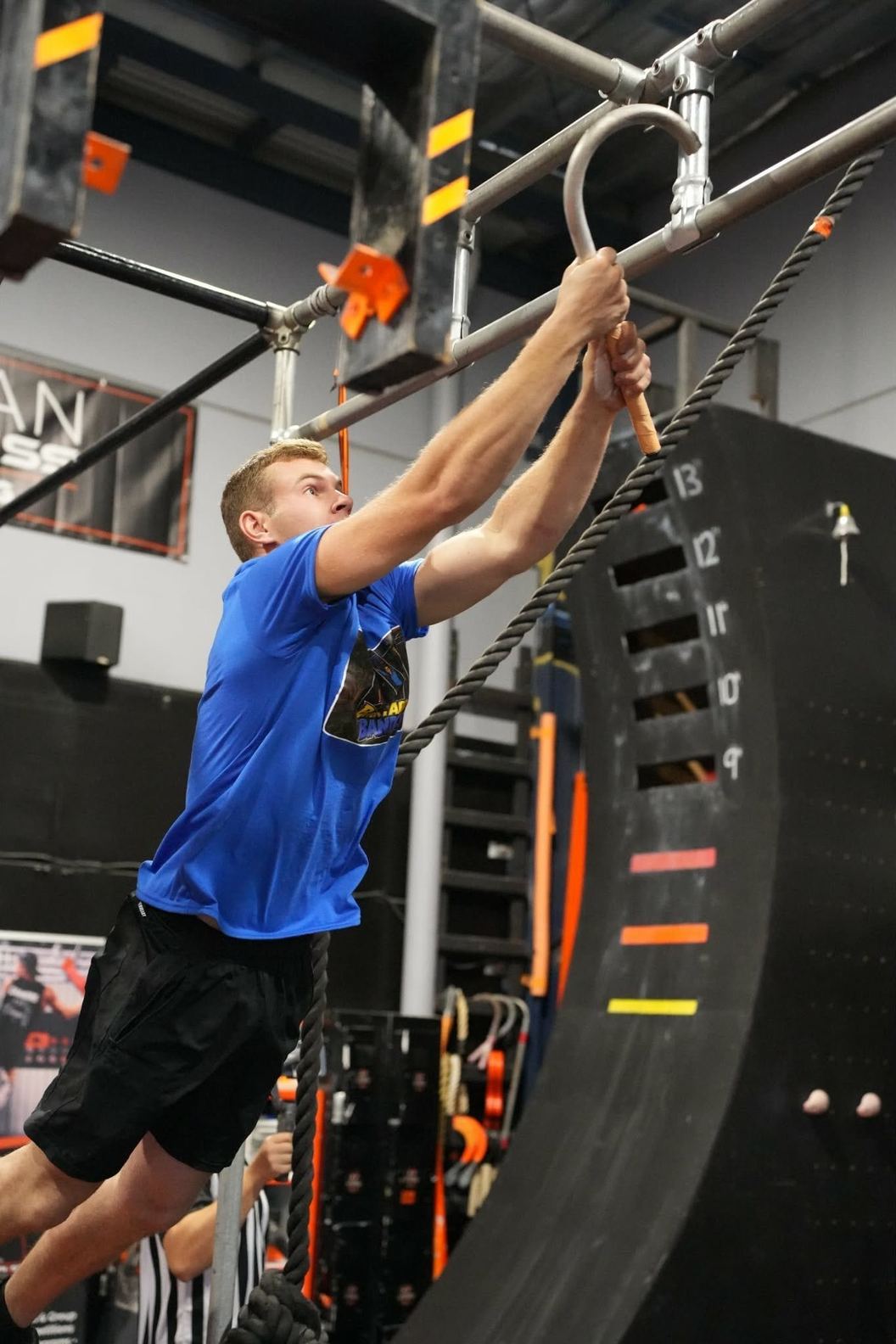 Athlete in a blue shirt pulling up on a hanging ring beside the AWF warped wall