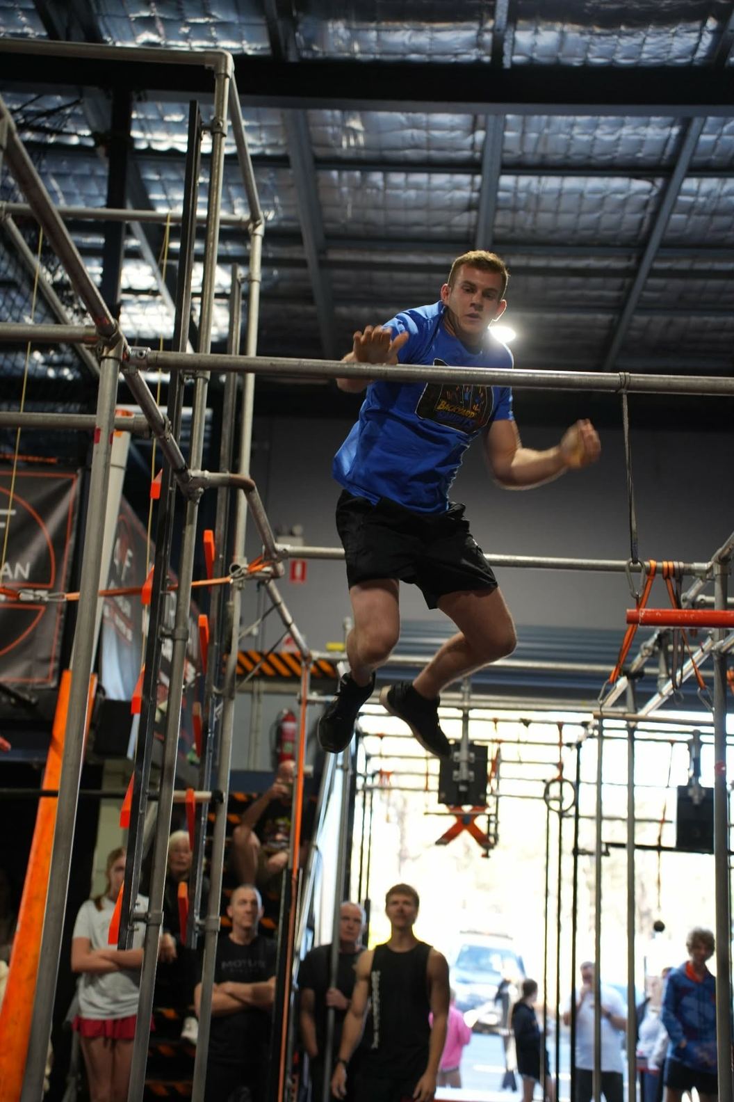 Athlete in blue leaping between bars on the AWF rig as a crowd looks on